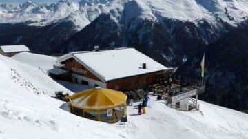 A mountain house in a snow-covered landscape with a view of the surrounding mountains. The terrace is equipped with a yellow sun umbrella and invites you to relax.
