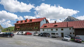 A large building with a red roof in a mountainous area. Several cars are parked in the parking lot, and the sky is blue with white clouds. | © TVB Paznaun - Ischgl