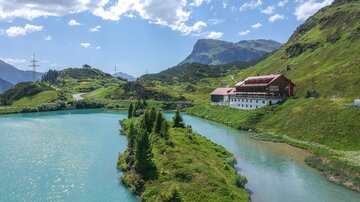 A picturesque landscape with a clear, turquoise lake and surrounding mountains. In the background, there is a traditional building amid green meadows. | © TVB Paznaun - Ischgl