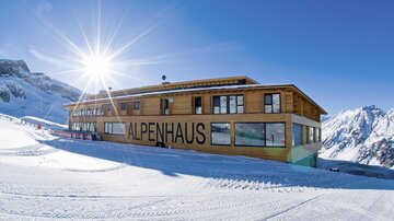 Ein modernes Holzgebäude mit der Aufschrift "ALPENHAUS" in einer schneebedeckten Berglandschaft. Die Sonne scheint strahlend am klaren blauen Himmel.