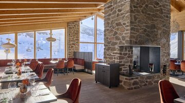 Ein modernes Restaurant mit großen Fenstern und Blick auf schneebedeckte Berge. Die Einrichtung besteht aus Holzelementen und gemütlichen, roten Stühlen.