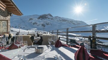 Eine gemütliche Restaurantterrasse mit Blick auf schneebedeckte Berge und strahlendem Sonnenschein. Der Tisch ist festlich gedeckt mit Gläsern und Servietten.