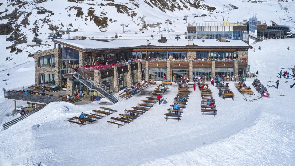 Ein modernes Berghaus umgeben von schneebedeckten Bergen. Viele Liegen stehen bereit, um die Sonne zu genießen.