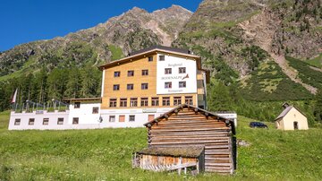 A modern building in a mountainous landscape. In the foreground stands a traditional wooden house.
