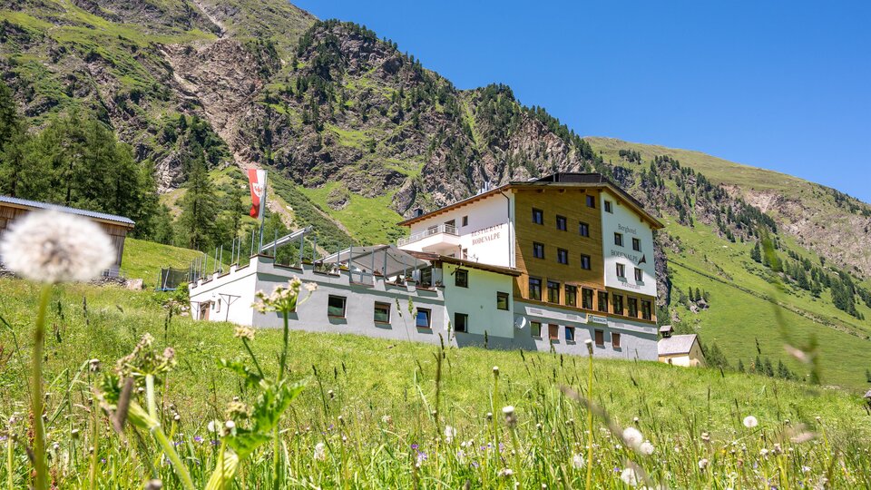 A large building in the mountains, surrounded by green meadows and rocks. The sky is clear and blue.