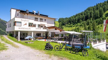 A modern mountain hotel with beautiful facade design. Bicycles are parked in front of the hotel and the green mountain forest stretches out in the background.