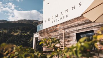 A modern restaurant facade with the inscription "Hermann's Kitchen". In the background, mountains and a blue sky are visible.
