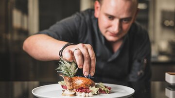 A chef decorates a carefully arranged dish on a white plate. The presentation includes fresh herbs and an appealing arrangement of ingredients.