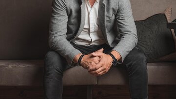 A smiling man is sitting on a couch in front of a wall with wines. He is wearing a gray blazer and white sneakers.