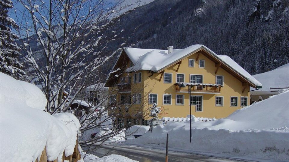 Ein charmantes, gelbes Haus im Schnee mit einem malerischen Berghintergrund. Der Weg ist von frischem Schnee bedeckt und die Bäume sind winterlich verziert.