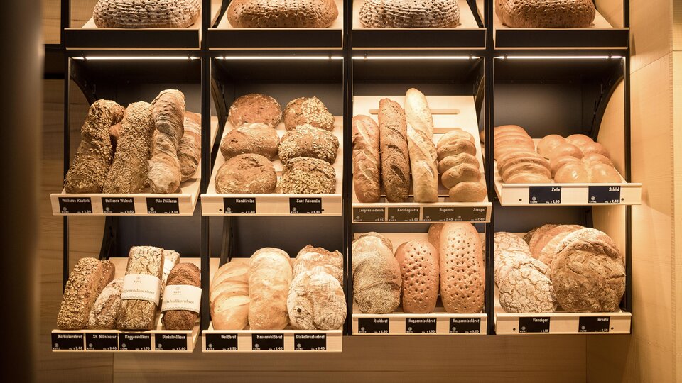 A selection of fresh bread in various shapes and colors. The breads are neatly arranged on shelves.