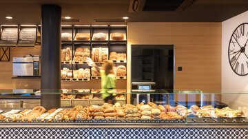 A modern bakery with a selection of fresh bread and pastries. In the background, an employee can be seen restocking the shelves.