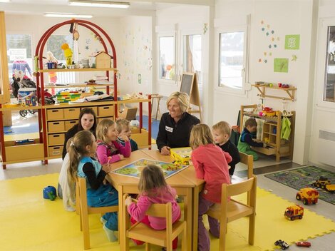 A bright kindergarten room with children sitting around a table and playing. A caregiver is present, while in the background, other children are occupied with toys.