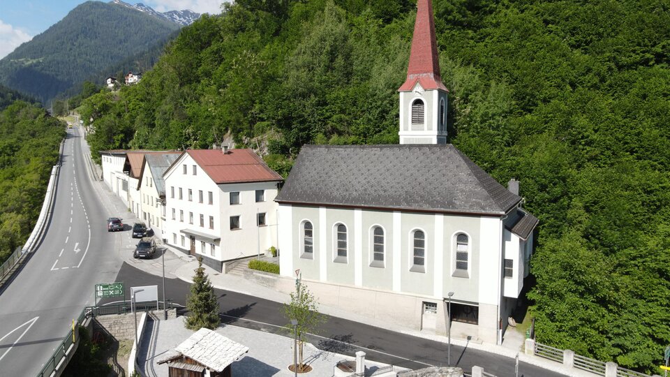 Eine kleine Kirche mit einem roten Dach und einem hohen Turm steht an einer Straße. Umgeben von grünen Hügeln und Wohngebäuden.