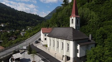 Eine malerische Kirche mit einem hohen roten Turm, umgeben von grünen Hügeln. Die Straße verläuft in der Nähe und es gibt einige Bäume und Gebäude in der Umgebung.