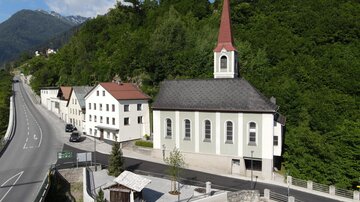 Eine kleine Kirche mit rotem Turm steht neben einer Straße, umgeben von grünen Bäumen. Im Hintergrund sind Berge sichtbar, und die Architektur ist typisch für eine ländliche Gegend.