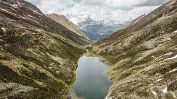 Eine malerische Berglandschaft mit einem klaren See zwischen hohen Bergen. Über dem Tal sind Wolken und die schneebedeckten Gipfel sind in der Ferne sichtbar.