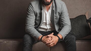 A smiling man is sitting on a couch in front of a wall with wines. He is wearing a gray blazer and white sneakers.