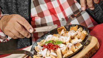 Ein Teller mit frisch zubereitetem Kaiserschmarrn, garniert mit Puderzucker und roten Johannisbeeren. Eine Person schneidet die süße Speise mit Messer und Gabel.