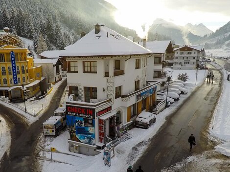 A charming building in the snow with a "CHECK IN" sign. In the background, there are snow-covered mountains and a quiet road.