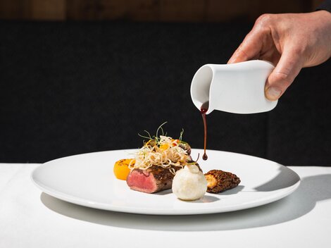 A stylish food presentation on a white plate. A hand is pouring sauce over the dish, which consists of meat, colorful vegetables, and side dishes.
