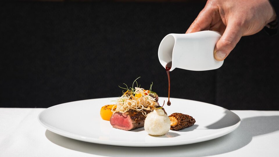 A stylish food presentation on a white plate. A hand is pouring sauce over the dish, which consists of meat, colorful vegetables, and side dishes.