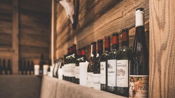 A row of wine bottles is on a wooden table. In the background, rustic walls can be seen.