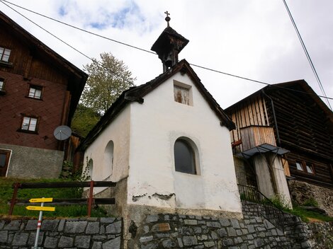 A small white chapel among traditional wooden houses. In the background, trees and a cloudy sky can be seen.