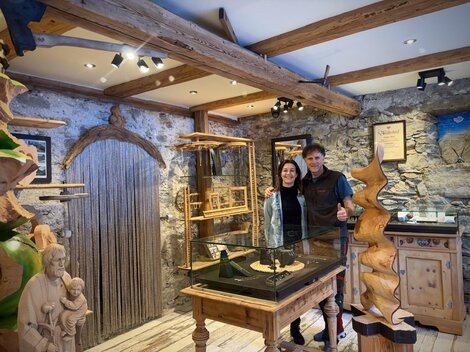 A woodworking shop with displayed sculptures and a workshop. Two people are standing in the middle of the room and smiling.