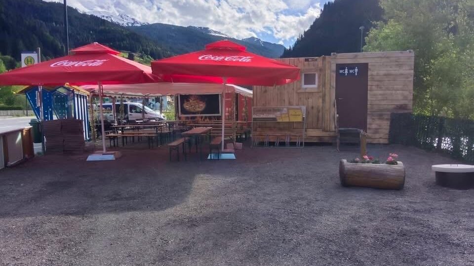 Ein kleiner Imbissstand mit Holzstruktur und roten Sonnenschirmen. Die Umgebung ist bergig und grün, ideal für eine Pause im Freien.