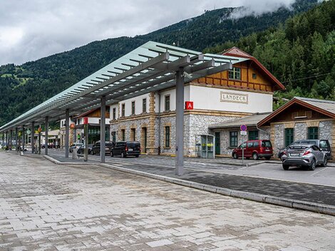 A train station building with traditional architecture and modern roofing. In the background, green mountains and clouds can be seen.