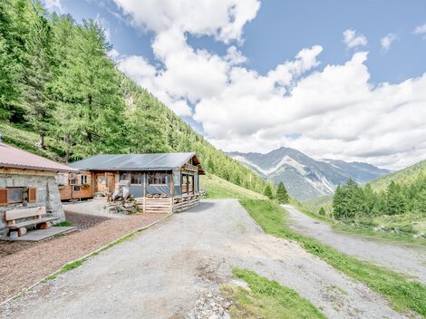 A picturesque Alpine landscape with two cottages and green trees. The mountains in the background are visible under a clear sky.