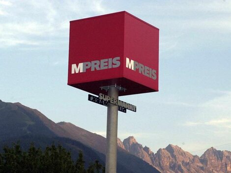 A red business sign with the inscription "MPREIS" in front of a mountain panorama. The sky is clear and the landscape is green.