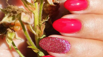 Bright red nails with glitter on a finger holding a plant with blackberries. The background shows green leaves.