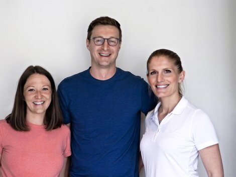 Three smiling individuals stand in front of a bright wall. They wear casual clothing and appear friendly and open.