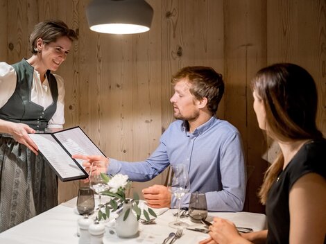 A waitress serves the menu to a couple in a cozy restaurant. The warm wooden walls create an inviting atmosphere.
