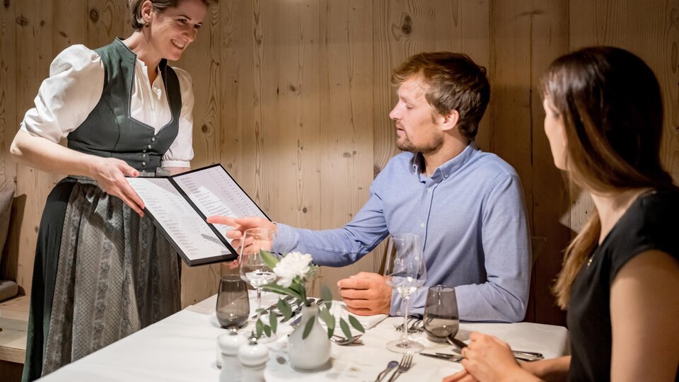 A waitress serves the menu to a couple in a cozy restaurant. The warm wooden walls create an inviting atmosphere.