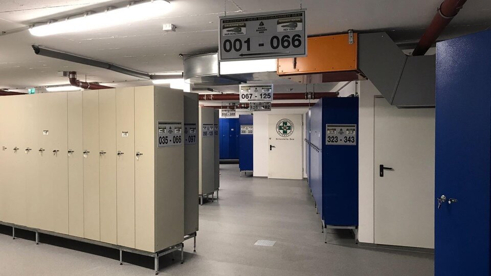 Ein moderner Umkleidebereich mit Spinden und geräumigen Gängen. Die Wände sind in Blau und Beige gehalten, und es gibt mehrere Nummernschilder.