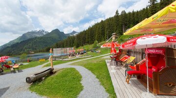 A beautiful viewpoint by the lake with deck chairs and umbrellas. In the background, mountains and forests are visible.