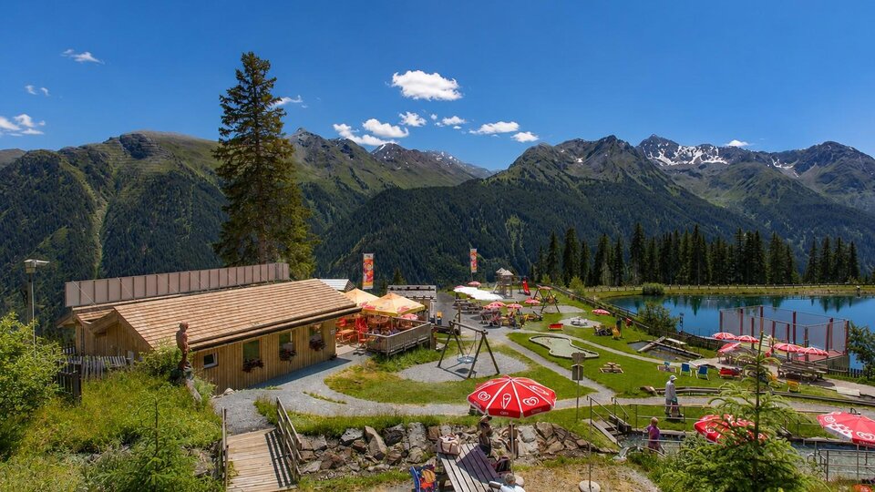 An alpine recreational facility set against an impressive mountain backdrop: In the foreground, a wooden terrace, sunbathing lawn, playground, and red sun umbrellas invite you to linger, while on the right, a clear mountain lake reflects the surrounding peaks. The bright wooden building, green meadows, and wide blue sky create a summery, family-friendly atmosphere in the heart of the Alps.
