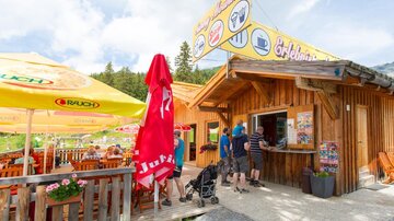 A cozy wooden snack stand in nature. There are tables with umbrellas and visitors are ordering food.