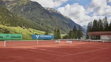 A tennis court with red sand, surrounded by picturesque mountains and green trees. The sky is cloudy, but the landscape is stunning.