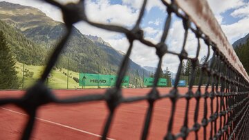A tennis court with a red surface, surrounded by mountains and trees. The net in the foreground reveals the playing surface.