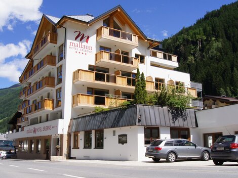 Ein modernes Hotelgebäude mit mehreren Etagen und Balkonen. Umgeben von grünen Bergen und klarem Himmel.