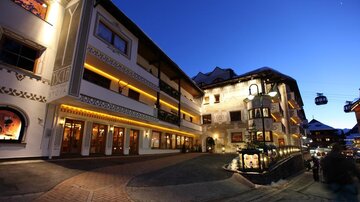 A charming alpine landscape at dusk with illuminated buildings. In the background, cable cars and snow-covered mountains can be seen.