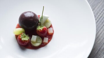 An elegant dessert on a white plate. It consists of a round type of chocolate and various fresh fruits.