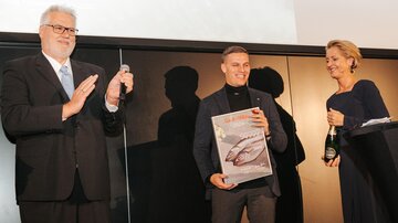 Three people are standing on a stage. The man in the middle is holding an award and is being applauded by the others.