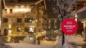 A picturesque winter village with snow-covered buildings and festive lighting. In the foreground, the logo of the Michelin Guide 2026 is visible.